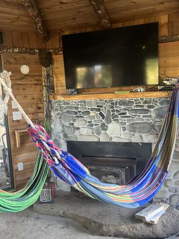 This cozy cabin room features a stone fireplace, a large TV mounted above, wooden walls and ceiling, colorful hammocks, and rustic accents for a relaxing vibe.