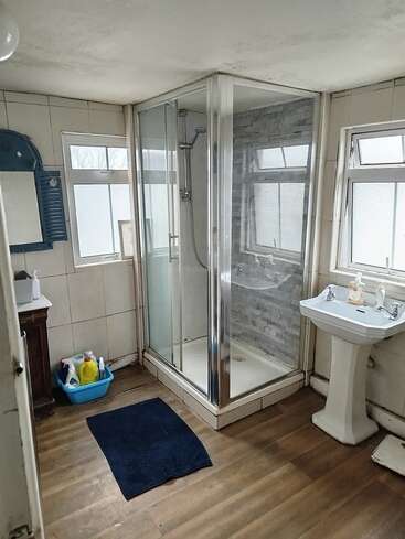 This bathroom features a glass shower, pedestal sink, blue mirror, cleaning supplies in a plastic tub, three frosted windows, tiled walls, and wooden flooring, creating a functional space.
