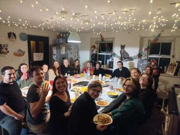 A group of people are gathered around a large table, enjoying a meal together in a festive, warmly-lit room decorated with string lights and cat pictures.
