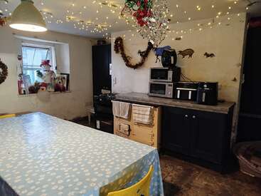 A cozy kitchen decorated for Christmas with fairy lights, garlands, a snowman on the windowsill, a long table with a blue floral tablecloth, and appliances.