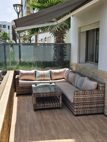 A cozy outdoor patio with a wicker L-shaped sofa, colorful pillows, glass-top coffee table, wooden floor, glass railing, palm trees, and a retractable awning.
