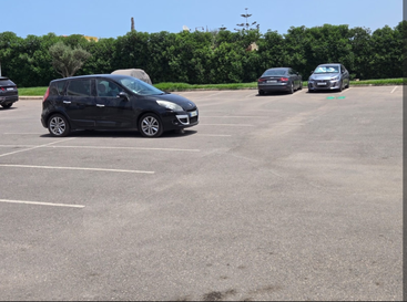 A mostly empty parking lot with a few parked cars, clear sky above, and green bushes forming the background along the lot’s edge. Sunny outdoor scene.