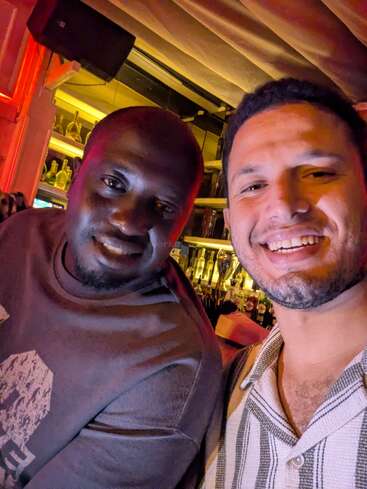 Deux hommes sourient et prennent un selfie ensemble dans un bar. L'arrière-plan est composé d'étagères avec des bouteilles et un éclairage ambiant chaleureux remplit l'espace.