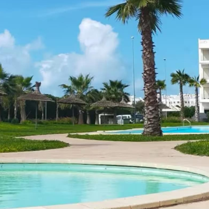 A sunny, tropical resort scene with a clear blue sky, palm trees, two pools, and thatched umbrellas. Modern white buildings are visible in the background. Relaxing atmosphere.