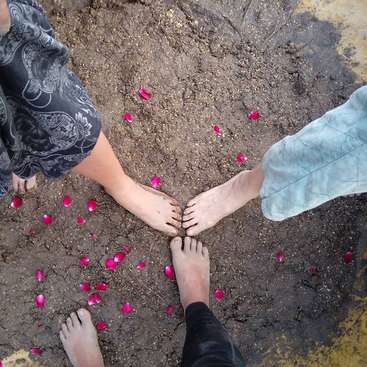 The image depicts four pairs of feet standing in the dirt, surrounded by rose petals, with each pair connected by hands. The scene is viewed from above.