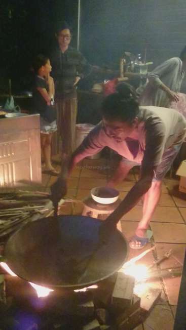 The image depicts a person stirring a large wok over an open flame, with a small child and two adults observing in the background.