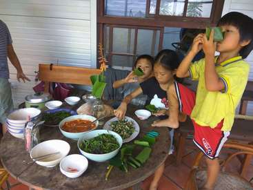The image depicts three children standing around a table laden with an assortment of dishes, including bowls of food and utensils, in a cozy outdoor setting.