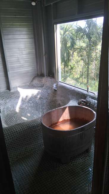 The image depicts a serene bathroom with a large window, featuring a wooden bathtub filled with water and a tiled floor, surrounded by lush greenery outside.
