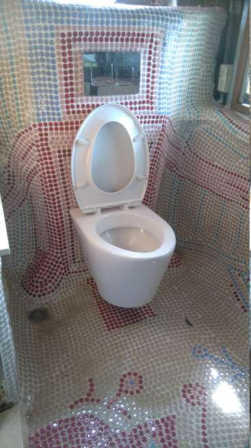 The image depicts a white toilet with an open lid, situated within a bathroom featuring a mosaic tile wall and floor, showcasing a vibrant and colorful design.