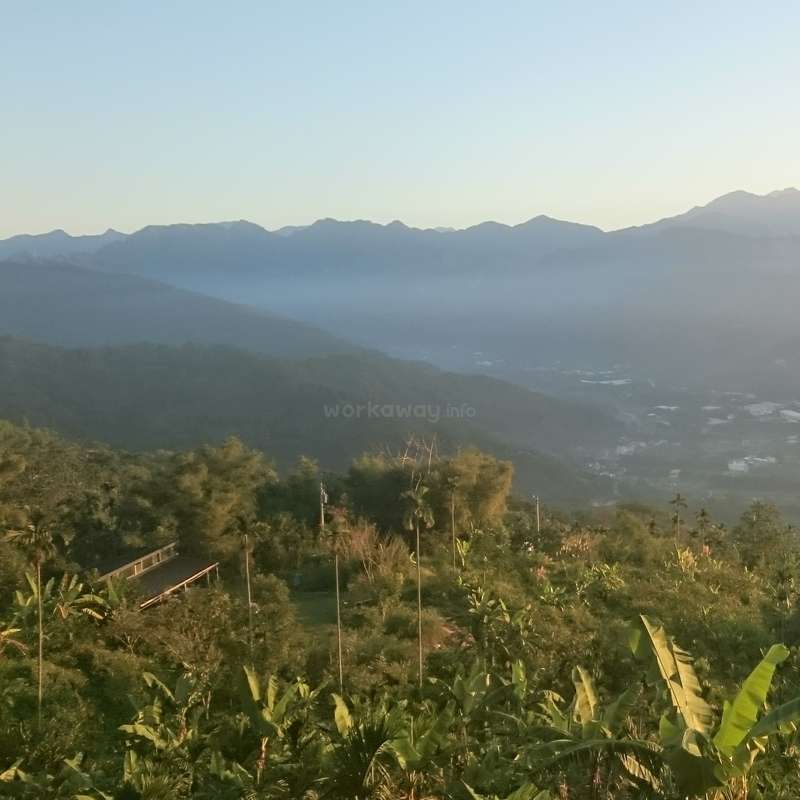 The image depicts a serene mountainous landscape with lush vegetation, a bridge, and distant mountains, set against a clear blue sky.
