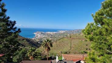 The image depicts a serene coastal town nestled in a lush valley, with a vibrant blue sky and a tranquil ocean in the background, surrounded by palm trees and green hills.