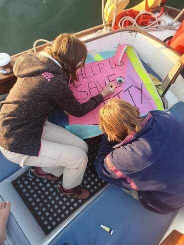 Zwei Personen auf einem Boot gestalten ein Schild mit der Aufschrift "HELP US SAVE..." mit schwarzem Marker auf einem rosa Stoff, möglicherweise für eine Notsituation.