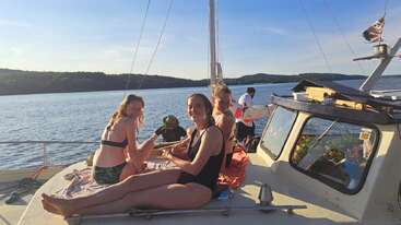 Vier Menschen entspannen und lächeln auf einem Boot unter sonnigem Himmel, mit ruhigem Wasser und fernen Hügeln im Hintergrund. Es sieht nach einem fröhlichen Sommertag aus.