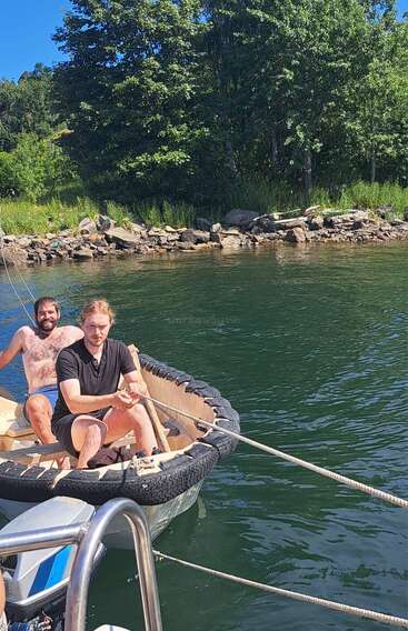 Zwei Männer sitzen in einem kleinen Boot, das aus einem Autoreifen gebaut wurde, und treiben auf einem ruhigen See in der Nähe eines felsigen, baumbestandenen Ufers unter sonnigem, blauem Himmel.