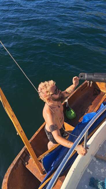 Ein hemdsärmeliger, tätowierter Mann mit blondem Haar und Sonnenbrille steht in einem kleinen Holzboot neben einem größeren Boot, umgeben von tiefblauem Wasser.