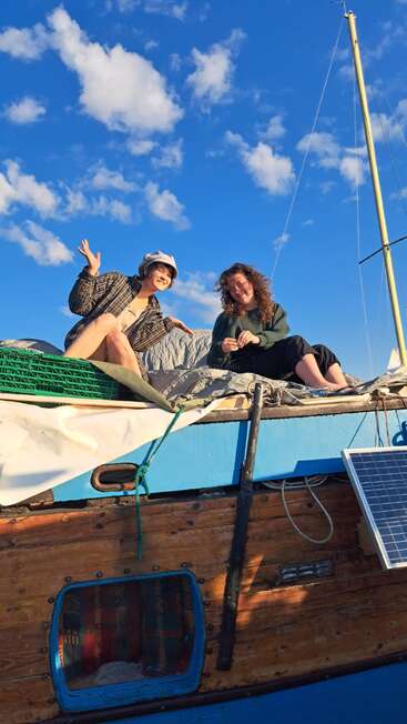Zwei Frauen sitzen lächelnd auf einem hölzernen Segelboot unter einem strahlend blauen Himmel mit flauschigen Wolken, genießen Sonnenschein und Entspannung und strahlen gemeinsam Freude und Abenteuer aus.
