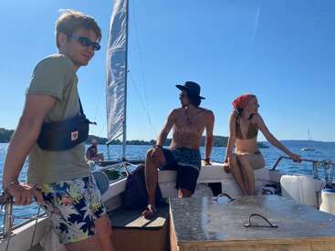 Drei Freunde entspannen sich auf einem Segelboot unter einem klaren blauen Himmel. Das Sonnenlicht spiegelt sich auf dem Wasser. Lässige Sommeroutfits und entspannte, glückliche Gesichtsausdrücke bestimmen die Szene.