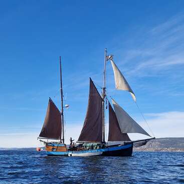 L'image représente un voilier à trois voiles sur un grand plan d'eau, avec pour toile de fond un ciel bleu parsemé de nuages et un rivage lointain.