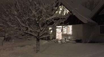 Ein gemütliches Haus leuchtet warm in der Nacht, umgeben von schwerem Schnee. Die kahlen Äste der Bäume sind mit Schnee bedeckt und schaffen eine friedliche, ruhige Winterszene im Freien.