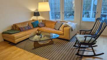 Im gemütlichen Wohnzimmer stehen ein hellbraunes Sektionssofa mit bunten Kissen, ein Schaukelstuhl, ein Couchtisch aus Glas, ein gemusterter Teppich und Stehlampen. Große Fenster.