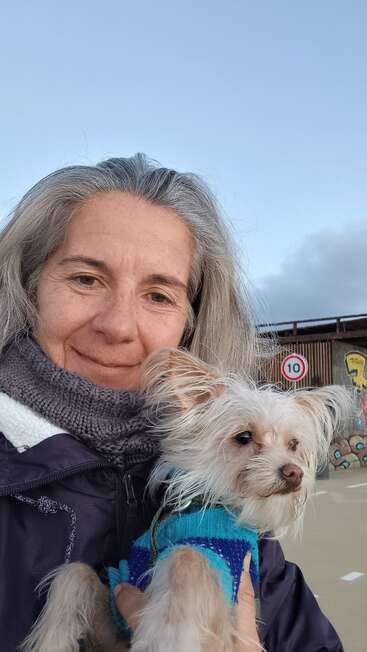 Eine Frau mit grauen Haaren und einem Schal lächelt, während sie einen kleinen Hund in einem blauen Pullover hält; sie sind draußen in der Nähe von Graffiti und einem 10-km/h-Schild.