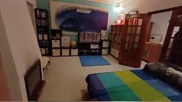 Ein gemütlicher, hell beleuchteter Raum mit Bücherregalen, einem bunten Bett, einem blauen Teppich, abstrakten Wandbildern, Holztüren und ordentlichen Aufbewahrungsboxen, der eine entspannte und einladende Atmosphäre schafft.