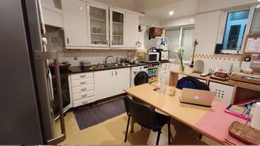 Diese gemütliche Küche hat weiße Schränke, einen Holztisch mit einem Laptop, Stühle, Geschirr neben der Spüle, einen Kühlschrank, Pflanzen und verschiedene Küchengeräte und -utensilien.