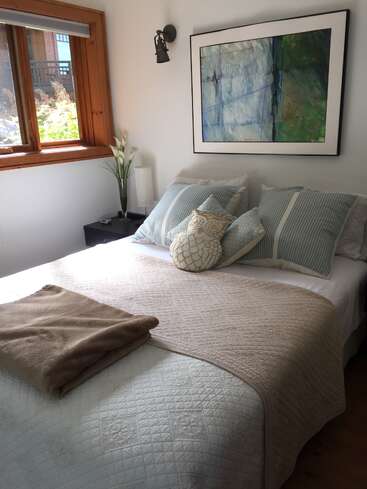 Das Bild zeigt ein ruhiges Schlafzimmer mit einem ordentlich gemachten Bett, einer weißen Bettdecke, blauen und weißen Kissen und einer beigen Decke vor einer strahlend weißen Wand.