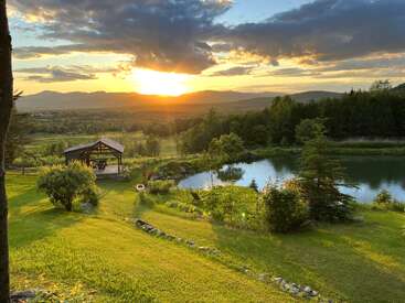 Das Bild zeigt eine ruhige Landschaft mit einem Teich, einem Pavillon, Bäumen und Bergen, die in das warme Licht eines Sonnenuntergangs getaucht sind.