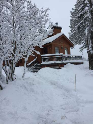 Das Bild zeigt ein gemütliches braunes Holzhaus mit schneebedeckter Veranda und Schornstein, eingebettet zwischen Bäumen, mit einem verschneiten Hof und einem Weg, der zur Haustür führt.