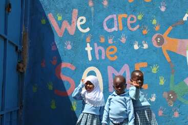 Tres niños con uniformes azules están de pie frente a un colorido mural en el que se lee «Somos IGUALES», rodeados de vibrantes huellas de manos, que simbolizan la unidad y la igualdad.