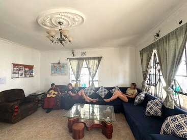 Três pessoas relaxam em uma sala de estar bem iluminada; uma toca violão enquanto as outras descansam em um sofá azul. A luz do sol entra pelas grandes janelas com cortinas.