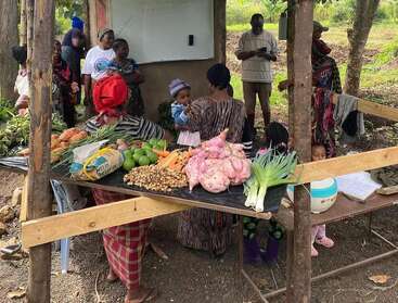 Un grupo de personas se reúne al aire libre alrededor de un puesto de mercado con verduras frescas, como ñames, zanahorias y verduras de hoja verde. Niños y adultos interactúan bajo los árboles.