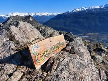 Un panneau en bois indiquant "Currumahuida" repose sur un terrain rocheux au sommet d'une montagne. Des pics enneigés, un ciel bleu clair et une vallée pittoresque offrent une vue à couper le souffle.