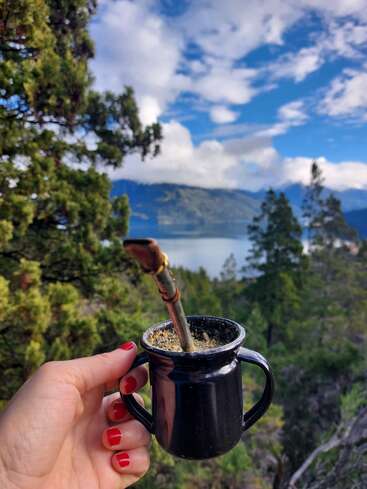 Une main aux ongles peints en rouge tient un gobelet de maté noir devant un paysage pittoresque composé d'arbres verts, d'un lac, de montagnes et d'un ciel partiellement nuageux.