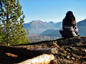 Une femme est assise sur une corniche rocheuse, contemplant des montagnes lointaines et un ciel bleu limpide. Des pins encadrent cette scène magnifique, paisible et naturelle.