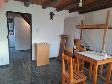 Um cômodo rústico com vigas de madeira expostas, uma pequena mesa com duas cadeiras, mobília mínima, piso de pedra e uma porta aberta que leva a outra área.