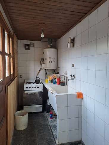 Uma cozinha estreita com paredes de azulejos brancos, teto de madeira e eletrodomésticos básicos. Você pode ver o fogão, a pia, o aquecedor de água, os utensílios, os materiais de limpeza e uma lixeira. Iluminação simples.