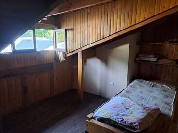Este é um quarto pequeno e rústico no sótão, com paredes e teto de madeira, teto inclinado, cama de solteiro, janela aberta, prateleiras e algumas roupas de cama dobradas. Ambiente aconchegante.