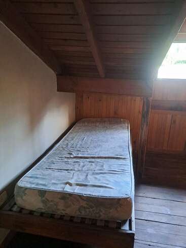 Esta imagem mostra um quarto simples de madeira no sótão com um único colchão velho em uma estrutura de cama, teto inclinado, painéis de madeira e luz natural de uma pequena janela.