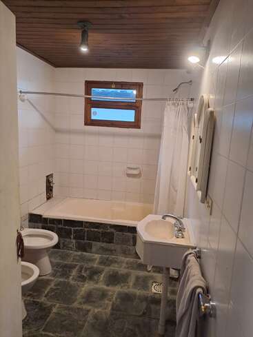 Esse banheiro tem uma banheira com cortina de chuveiro, bidê, vaso sanitário, pia pequena, piso de pedra, teto de madeira, paredes de azulejos brancos e uma janela.