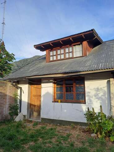 Uma pequena casa com telhado de metal corrugado, janelas e porta de madeira. Cercada por grama, plantas, parede de tijolos e montanhas visíveis ao fundo.