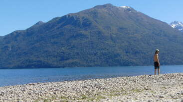 Um homem está em uma praia rochosa à beira de um lago límpido, cercado por montanhas com manchas de neve, sob um céu azul brilhante e sem nuvens.