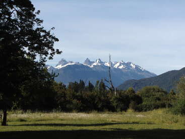A imagem mostra uma paisagem cênica com campos gramados, árvores espalhadas e floresta densa. Majestosas montanhas cobertas de neve surgem ao fundo sob um céu claro.