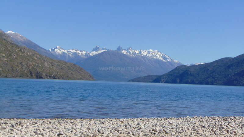 A serene blue lake is surrounded by forested mountains with snow-capped peaks. The shore is covered with small white stones, underneath a clear blue sky.