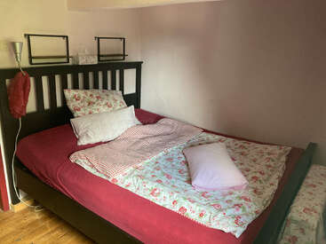 Uma cama aconchegante com roupa de cama com padrão floral, lençol vermelho e almofadas brancas. Cabeceira de madeira, candeeiro de leitura e prateleiras fixadas numa parede clara e simples, piso de madeira.