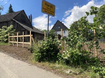 Cette image représente un paysage rural avec un panneau indiquant la localité de « Hardt », une clôture en bois, des buissons verts, une vieille grange, une maison et un ciel partiellement nuageux.