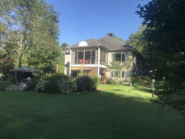 A large two-story house sits amid lush greenery, gardens, and tall trees. There's a gazebo on the lawn, under a bright blue sky. Peaceful.