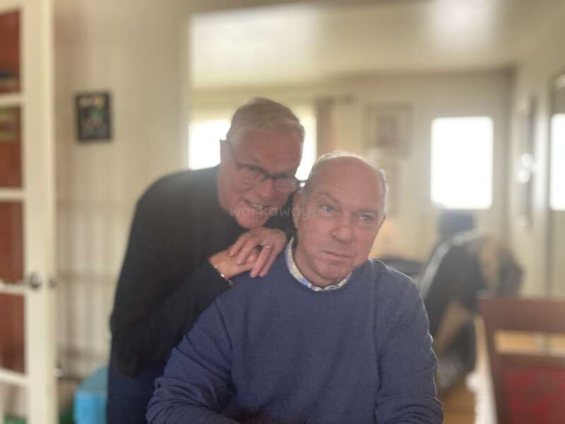 Two elderly men are indoors. One is sitting and the other is standing behind, hands resting on his shoulders. The room is warmly lit and cozy.