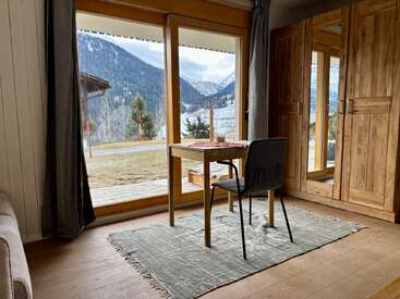 Ein gemütliches Bergzimmer verfügt über einen kleinen Tisch und einen Stuhl neben großen Fenstern, die einen atemberaubenden Blick auf schneebedeckte Gipfel bieten, sowie über Holzmöbel und viel Tageslicht.